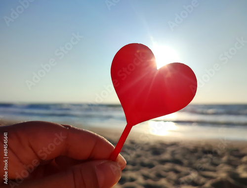 Person holding in fingers hand stick in shape red heart on background sea and sea waves, blue sky in seashore on sunny summer day close-up. Concept love romance amour St Valentines Day