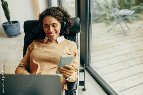 Smiling female freelancer working on laptop from home while use mobile phone sitting in chair