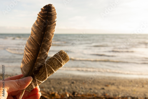 A tranquil image of a hand holding a white sage smudge stick with sacred feather with the Pacific Ocean in the background. 