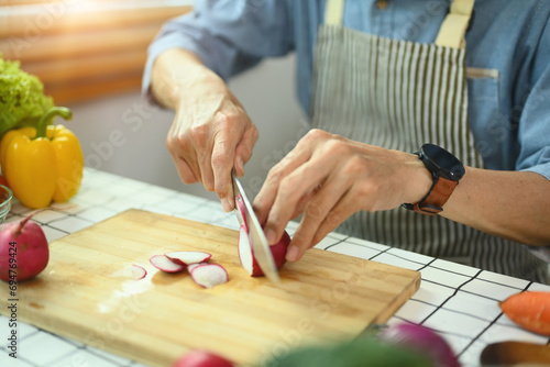 Senior man wearing apron cooking healthy food at home, slicing fresh radishes on cutting board.