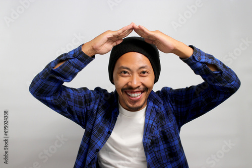 Happy Young Asian man with a beanie hat and casual clothes is making a hand gesture above his head, symbolizing a roof or home while standing against white background