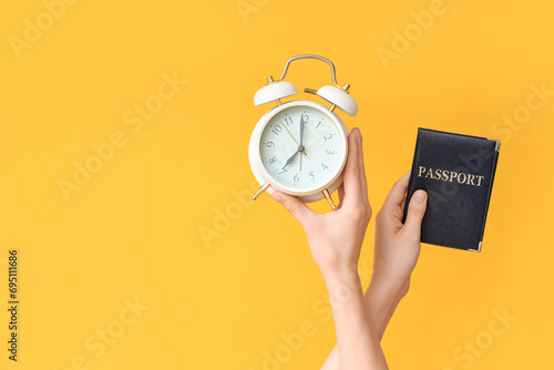 Female hands holding alarm clock and passport on yellow background