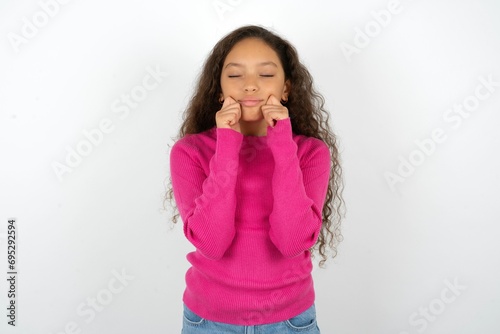 Pleased Beautiful teen girl wearing pink sweater over white background with closed eyes keeps hands near cheeks and smiles tenderly imagines something very pleasant