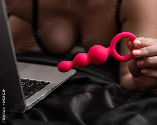 Woman holding pink anal beads next to laptop while lying on black sheet. 