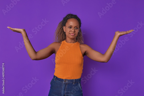 Young pragmatic ethnic African American woman spreads arms to sides showing scales and offering to compare two product options to make best choice stands on isolated purple background.