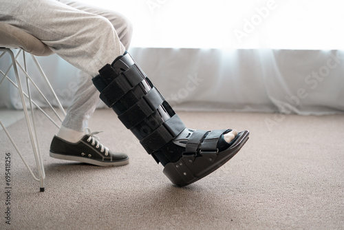 Foot orthosis concept. Close-up of man leg with ankle orthosis, black splint, walker boot on the sofa, profile view.