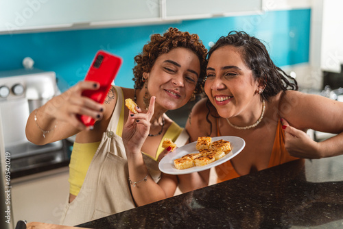 Amigas tirando uma selfie na cozinha, segurando o prato que prepararam juntas.