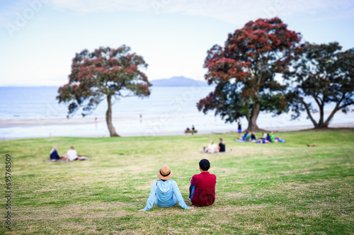 Couple relaxing on Takapuna beach. Pohutukawa trees in full bloom. Rangitoto Island in the distance. Auckland.
