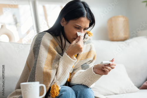 Tired woman with a cold taking her fever while lying with a blanket on the couch at home