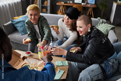 High angle view at diverse group of smiling young adults in living room holding slices of pizza and toasting with soda