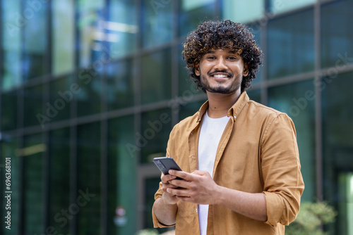 Portrait of a happy and smiling young Muslim man standing on a city street using a mobile phone and looking confidently at the camera