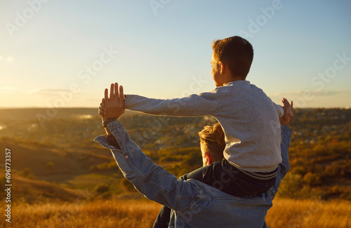 Fathers day.Close up portrait of a happy smiling son sitting on fathers shoulders and looking into the distance enjoying sunset and nature during a walk outdoors. Love, and family leisure concept.