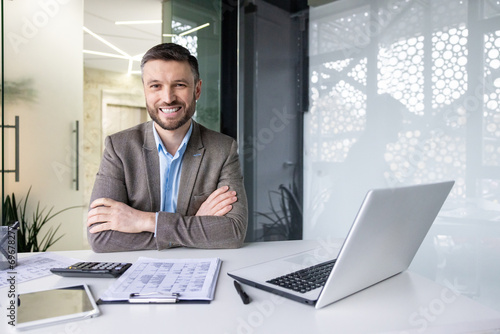 Portrait of successful financier accountant at workplace inside office, senior experienced businessman smiling looking at camera with crossed arms, working with papers sitting at table with laptop.