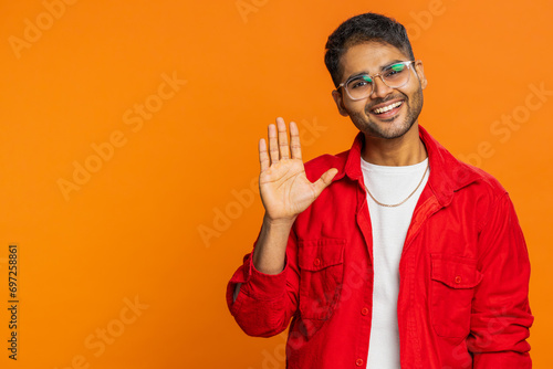 Young Indian man smiling friendly at camera, waving hands gesturing hello greeting, goodbye welcoming with invitation hospitable expression. Arabian Hindu guy isolated on orange background. Copy-space