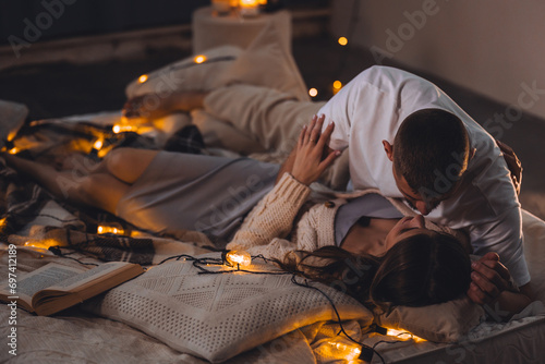 Beautiful happy loving young smiling couple relaxing in bed, watching movies on projector. Cozy home atmosphere, tenderness, closeness. Romantic surprise, atmospheric candles, Saint Valentine's Day