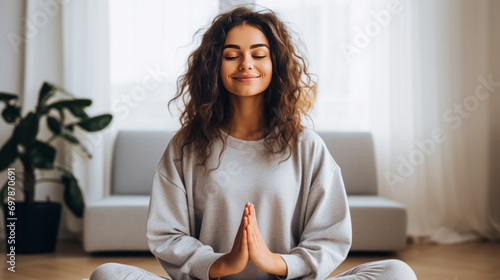 A content woman meditates with eyes closed, hands in prayer position in a cozy room.