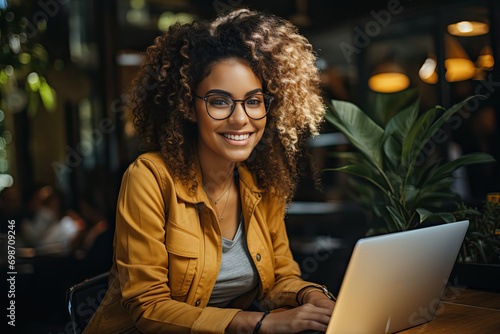 Joyful happy Black business woman in glasses sitting at workplace with laptop in home office, looking at camera, smiling. Millennial worker, employee, entrepreneur head shot portrait
