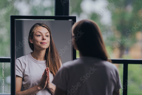 Reflective young woman in a casual t-shirt practicing self-affirmation in front of a mirror in a well-lit room.