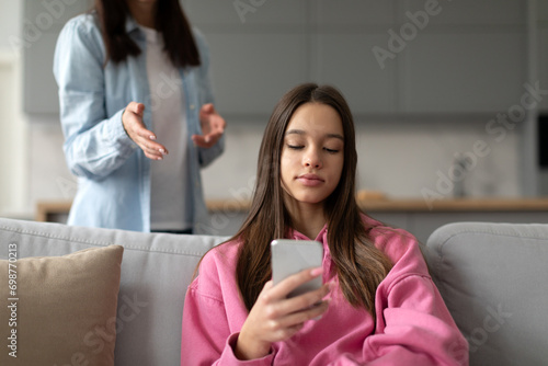 Angry mother having trouble with teenager girl, sitting on sofa at home and playing video games on smartphone