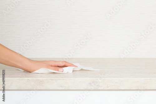 Young adult woman hand holding dry white paper napkin and wiping beige stone tabletop at home kitchen. Closeup. Cleaning service. Front view. Empty place for text on wallpaper background.