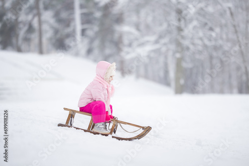 Happy little girl in pink warm clothes sitting on sledge and sledding down on snow from hill. Child enjoying white winter day at park. Side view.