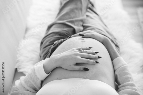 Cropped black and white photo taken from above of pregnant woman in unbuttoned blue jeans laying on white couch with hands on her bare pregnant belly aesthetic
