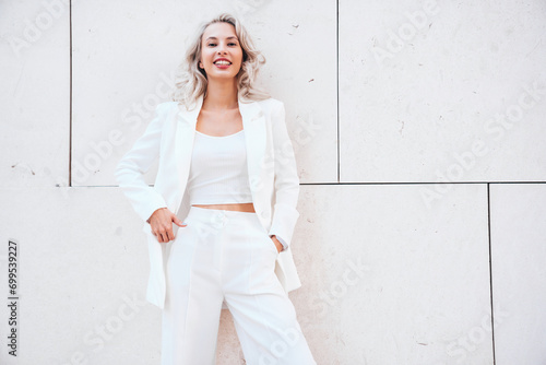 Young beautiful blond woman wearing nice trendy white suit jacket. Smiling model posing in the street at sunny day. Fashionable female outdoors. Cheerful and happy. posing near wall