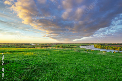 A river against a backdrop of green fields, a breathtaking view from the hill. Colorful sky with clouds during sunset. Landscape in spring. Nature during a bright sunset.