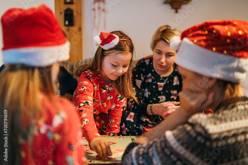 Mother with daughters kids playing together in table board game, wearing Christmas hats. Cozy pre Christmas evening time moment. Family values, board games and X-mas celebration concept.