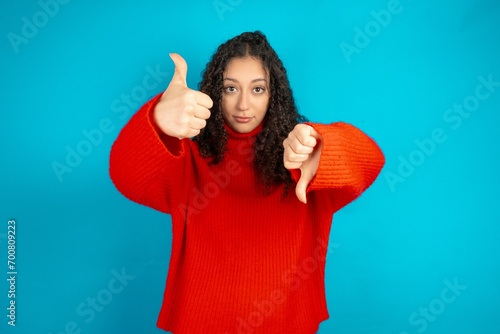 Beautiful teen girl wearing knitted red sweater over blue background showing thumbs up and thumbs down, difficult choose concept