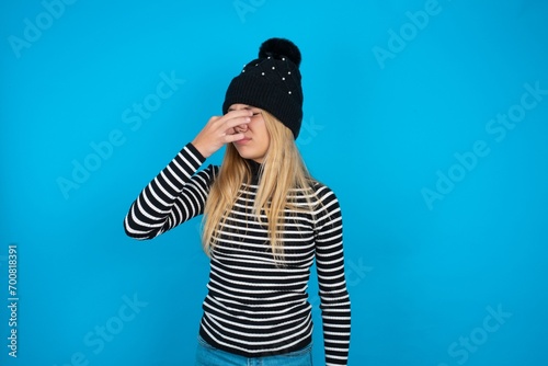 Teen caucasian girl wearing striped sweater and woolly hat smelling something stinky and disgusting, intolerable smell, holding breath with fingers on nose. Bad smell