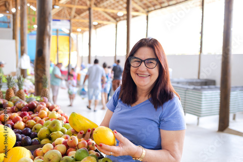A joyful and smiling brazilian mature woman at a market, holding two papayas, illustrates the everyday shopping experience on Consumer Day