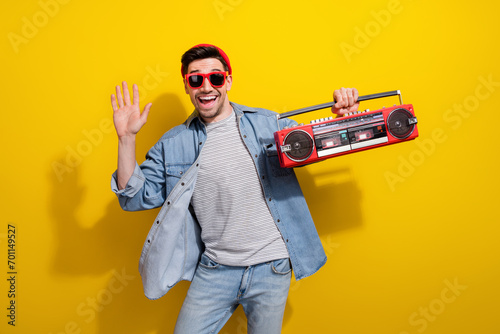 Photo of funky friendly man dressed jeans shirt dark spectacles waving arm hi listening boom box songs isolated yellow color background