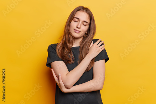 Selfish long haired woman with closed eyes embracing herself tenderly daydreams about something wears casual black t shirt isolated over yellow wall. Romantic female model recalls nice lovely memories
