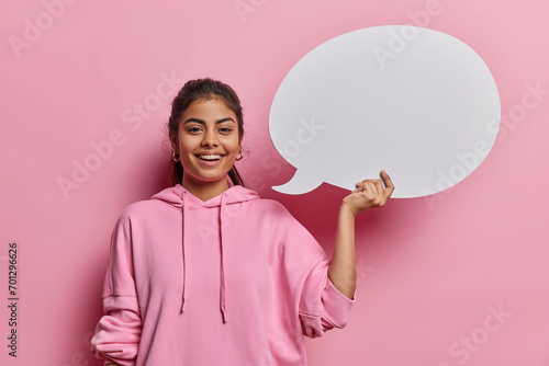 Positive lovely Iranian woman with dark hair smiles gladfully holds blank speech bubble gives promotional opportunity smiles gladfully wears casual sweatshirt poses against pink studio background.