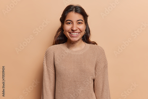 Portrait of pleasant looking Iranian girl with toothy smile has good mood dressed in casual knitted jumper looks directly at camera isolated over brown background. People and positive emotions concept