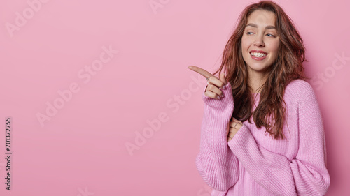 Smiling European woman with long dark wavy hair points index finger aside on copy space wears casual loose knitted jumper shows something pleasant isolated over pink background. Advertisement concept