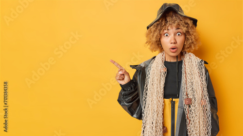 Indoor shot of impressed fisherwoman showcases her expertise poses with net around neck has fishing expedition wears black hat and leather jacket points index finger on empty space for your text