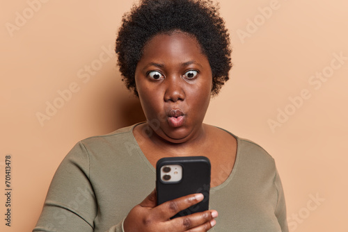Photo of stupefied dark skinned woman checkes mailbox reads disturbing message received fascinating offer holds smartphone feels impressed wears casual jumper isolated over brown background.