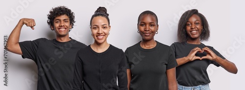 Horizontal shot of three diverse young women and one Hindu man smile pleasantly show heart gesture and raise arm demonstrates biceps wear same black t shirts isolated over white studio background.