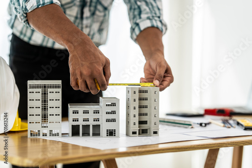 Close up Asian individual architect or engineer holding building scale model of multi-story building. design or review of architectural plans laid out on work surface in front of them.