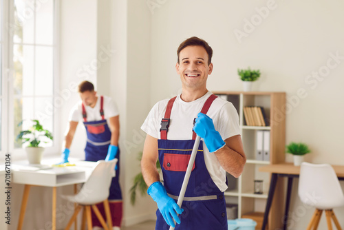 Young male cleaner in overall, happy smiling man performing cleaning duties, professional team busy sanitizing, sweeping, polishing, mopping floor, house cleaning services for home, office