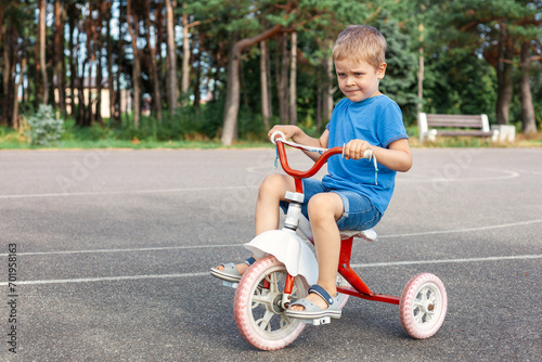 A cute very satisfied child is driving his old tricycle in the city outdoor basketball court