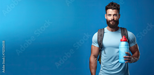 Muscular man with a bottle of water or isotonic drink in his hand on a blue background. The concept of sports nutrition and recovery after training in the gym. Empty space for product placement.