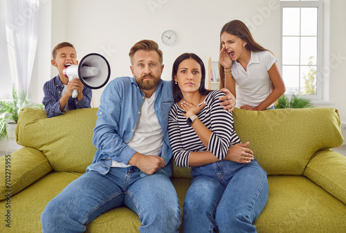 Children screaming at parents. Two naughty kids making lots of noise. Unruly brother and sister with megaphone screaming at mum and dad sitting on sofa in living room with scared face expressions