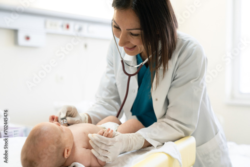 Smiling female pediatrician using stethoscope and protective gloves to check up a newborn baby.
