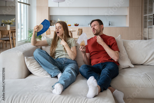 Suffering from heat family couple sitting on couch waving paper fans at home. Tired exhausted man woman feel bad in hot summer weather in room without air conditioner. Overheating, sultriness concept.