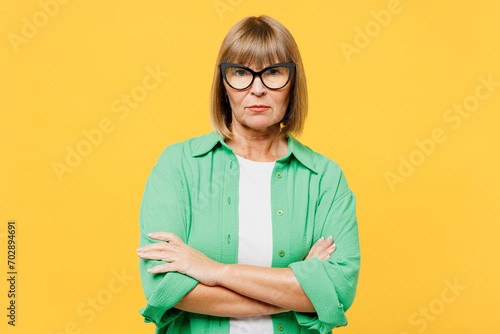 Elderly sad frowning blonde woman 50s years old wears green shirt glasses casual clothes hold hands crossed folded look camera isolated on plain yellow background studio portrait. Lifestyle concept.