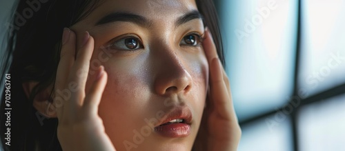 Asian girl examines her face in the mirror, concerned about skin condition, seeks cosmetology services for moisturizing and care.