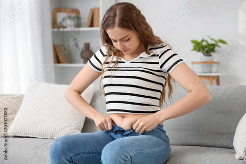 Young woman in tight jeans sitting on sofa at home. Weight gain concept